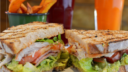A grilled sandwich packed with shredded turkey, fresh lettuce, and tomato slices is served on a plate. Behind the sandwich, there is a bowl of sweet potato fries, a glass of red drink, and a glass of orange drink.