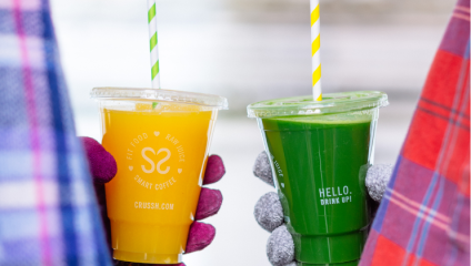 Two people holding beverages in plastic cups with striped straws. One cup contains an orange drink labeled FIT FOOD RAW JUICE SMART COFFEE, and the other has a green drink labeled HELLO. DRINK UP! Both individuals are wearing colorful, patterned clothing.