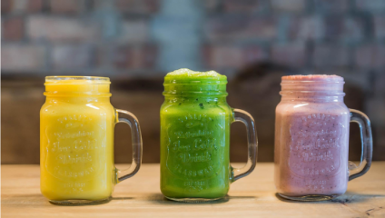 Three mason jars are lined up on a wooden surface, each filled with a different smoothie. From left to right: a yellow smoothie, a green smoothie, and a pink smoothie. The background is out of focus, with a mix of earthy tones.