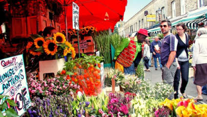 A colorful market scene with various flowers on display, including sunflowers, tulips, and Chinese lanterns. People walk and browse under bright red umbrellas. A man in a green and red outfit looks at the flowers, while others carry drinks and chat.