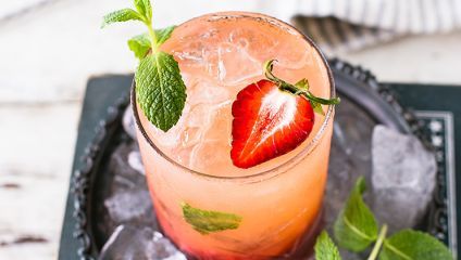 A refreshing drink served in a glass filled with ice. The beverage is light pink and garnished with a strawberry slice and a sprig of mint. The glass is on a metal tray with scattered ice cubes and a few mint leaves around it.