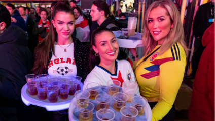 Three women are smiling and holding trays with plastic cups filled with drinks. They are standing in a crowd and each woman is wearing a different football jersey: England, Colombia, and possibly a third team. The atmosphere appears festive and lively.