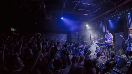 A lively crowd watches a band performing on a dimly lit stage. The band includes a keyboardist and guitarist under blue and purple stage lights. Audience members have their hands raised, enjoying the energetic atmosphere in the compact indoor venue.