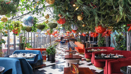 A vibrant restaurant space with a checkered black and white floor. The ceiling is adorned with hanging plants and colorful ornaments. The seating features a mix of red and blue velvet chairs and booths, with intricately set tables ready for diners.