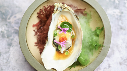 A beautifully plated dish featuring a single oyster, garnished with vibrant green and orange spheres, small purple flowers, and a green leaf. The oyster is placed on a bed of red and green seaweed, all presented on a light grey plate.