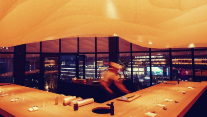 Spacious modern restaurant at night, featuring a sleek wooden table setup, minimalist chairs, and large floor-to-ceiling windows revealing a twinkling cityscape outside. Soft, curved ceiling decorations add an artistic touch. A blurred, moving figure is seen.