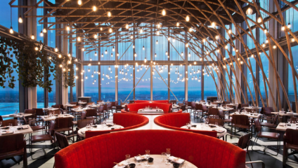 A modern restaurant with floor-to-ceiling windows overlooking a cityscape at dusk. The interior features exposed beams, hanging pendant lights, and red semicircular booths. Tables and chairs are neatly arranged around the spacious dining area.