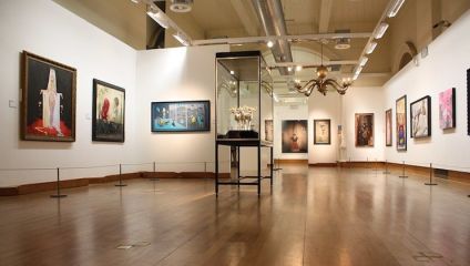A well-lit art gallery features various paintings and a central glass display case containing sculptures. Artwork includes portraits, abstract, and modern pieces, all evenly spaced on white walls. The wooden floor contrasts with the white walls, creating a warm ambiance.