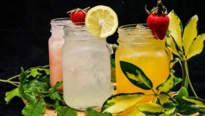 Three mason jar drinks are displayed with garnishes. The left jar is pink with a strawberry garnish, the middle jar is clear with a lemon slice, and the right jar is orange with a strawberry. Green leaves surround the jars, set against a black background.