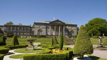A grand, historic mansion with a neoclassical facade and columns stands under a clear blue sky. The foreground features immaculately landscaped gardens with neatly trimmed hedges, colorful flowerbeds, and stone pathways, creating an elegant and serene atmosphere.
