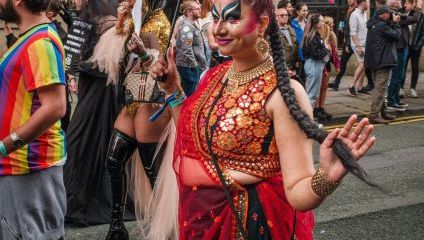 A person in vibrant, traditional attire and dramatic makeup walks under a pink and blue umbrella. They wear a red and gold sari with intricate patterns, and a beaded necklace, and carry a small colorful bag. Onlookers in casual clothing are in the background.