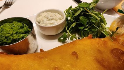 A plate of food featuring a large piece of battered and fried fish, accompanied by a small dish of green mushy peas, a small cup of tartar sauce, and a side of fresh green salad leaves. Part of a lemon wedge wrapped in a cloth is also visible on the plate.