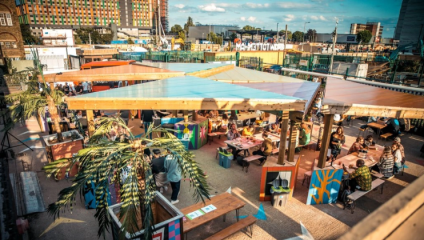 An outdoor venue with colorful canopy roofs and tables filled with people enjoying food and drinks. The area is decorated with potted palm trees and vibrant murals. In the background, there are multicolored buildings and a blue sky with scattered clouds.