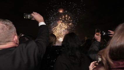 A group of people watches a vibrant fireworks display at night. Some are capturing the moment with their phones, while others hold glasses. The sky is illuminated with bursts of colorful fireworks.