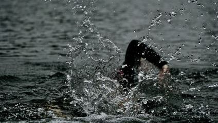 A swimmer in a black wetsuit is partially submerged in the water, with their hand reaching forward and water splashing around them. The splashes create a dramatic effect against the dark, rippling surface of the water.