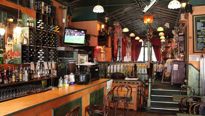 A cozy, dimly-lit bar with a wooden counter and stools. Shelves lined with bottles and glasses are behind the bar. A TV displaying a soccer match is on the wall. Tables and chairs are scattered around, with lantern-style lights and decorative elements creating a warm atmosphere.