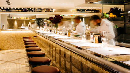 Inside a modern, upscale bar with marble counters, several chefs are seen in motion, preparing food behind a well-lit counter. The counter is lined with empty barstools and various condiments. Bottles and glassware are arranged neatly on shelves above.