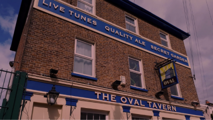A three-story brick building with blue accents houses The Oval Tavern. A sign with the words Live Tunes, Quality Ale, Secret Garden is displayed prominently at the top. A hanging pub sign shows the establishment's name and image of the building.