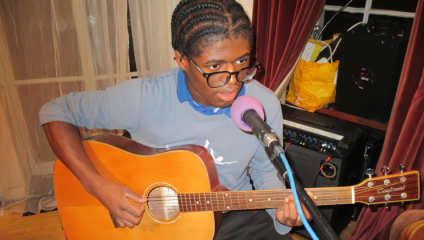 A person with braided hair and glasses strums an acoustic guitar, seated in front of a microphone. They are wearing a blue sweater and appear to be performing indoors, with curtains and various equipment visible in the background.
