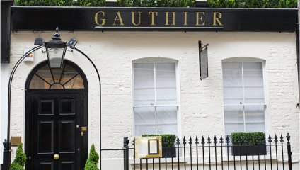 The façade of a restaurant named Gauthier featuring a white-painted brick exterior. It has a black door with an arched light fixture above it and two white-framed windows with white curtains. A black sign with gold lettering displays the restaurant's name.