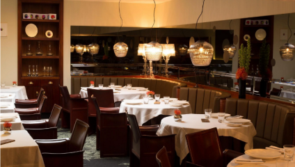 A cozy restaurant interior with elegantly set tables covered in white tablecloths. Wooden chairs surround the tables, and decorative light fixtures hang from the ceiling, casting a warm glow. A large mirror reflects the dining area, enhancing the ambiance.