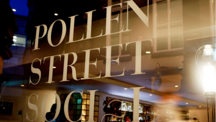 Close-up of a window with the words POLLEN STREET SOCIAL in large white letters. The reflection shows a cozy interior featuring blurred light fixtures and part of a bar area with bottles and glassware on shelves. The overall scene has a warm, inviting atmosphere.