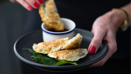 A pair of hands hold a dark plate with two pan-fried dumplings on a bed of green leaves. The left hand dips a dumpling into a small bowl of soy sauce, which is also on the plate. The person has red nail polish and wears a gold bracelet.