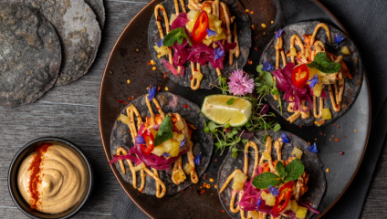 A plate with four gourmet tacos on blue corn tortillas, garnished with vibrant toppings such as sliced red chilies, pickled onions, fresh herbs, and edible flowers. A lime wedge and a bowl of creamy dip sprinkled with chili powder are beside the plate.