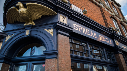 Storefront of the Spread Eagle pub, featuring a dark blue and gold color scheme. A golden eagle statue is perched above the entrance, and a sign displays the name Spread Eagle in gold letters. The building has a brick facade with a streetlamp visible.