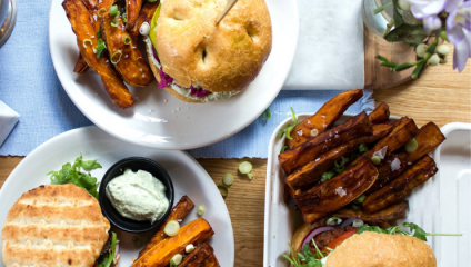 A top-down view of a table set with plates of food. Two sandwiches, one on a burger bun and another on a sandwich grill bread, are served with sweet potato fries and a side of dip. A small vase with flowers is partially visible on the upper right of the table.