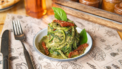 A round plate with zucchini noodles topped with basil pesto, sun-dried tomatoes, and fresh basil leaves is on a table. A fork and knife are placed on the table, along with a wooden spice holder and a bottle of oil in the background.