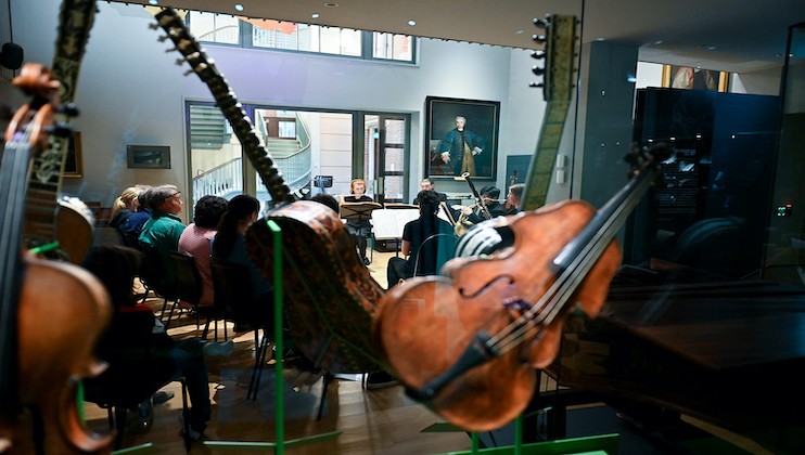 People attend a live classical music performance in a room, which is viewed through a display case containing string instruments like violins and cellos. The audience is seated, focusing on the musicians. A large portrait hangs on the wall in the background.