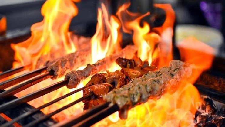 Skewers of various meats are being grilled over an open flame, with the fire providing a vibrant display of orange and yellow hues. The scene suggests a barbecue or cookout setting, capturing the intense heat and smoky atmosphere of the cooking process.