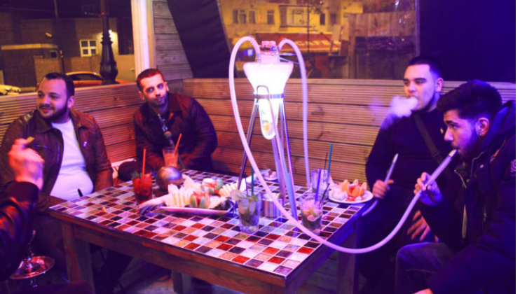 Five people are sitting around a table in a dimly lit place, possibly a lounge or café. The table has mosaic tiles and several drinks on it. A large hookah with multiple pipes is placed at the center, and one person is blowing out smoke.