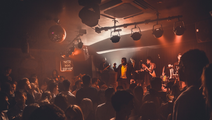 A lively crowd enjoys a concert in a dimly lit venue. On stage, a band performs energetically with a singer at the forefront. Spotlights illuminate the stage, and a disco ball hangs from the ceiling, adding to the vibrant atmosphere.