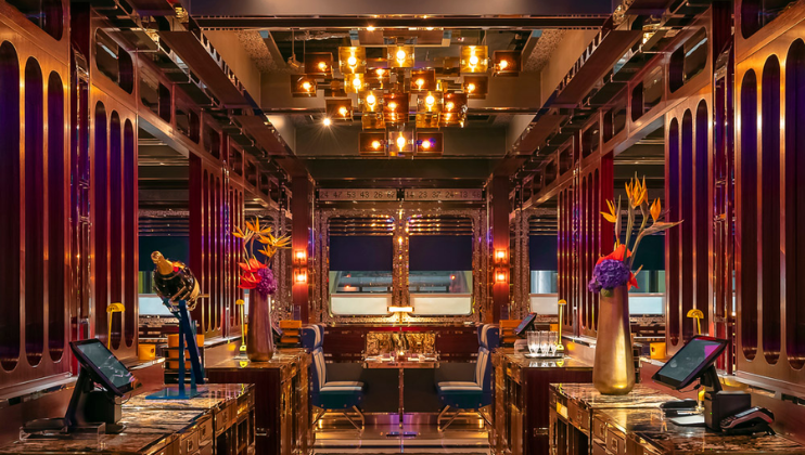 Luxurious restaurant interior with warm lighting, ornate wooden panels, and a central chandelier. Tables are set with white tablecloths, and floral arrangements adorn the space. A mannequin dressed in a suit adds a stylish touch in the foreground.