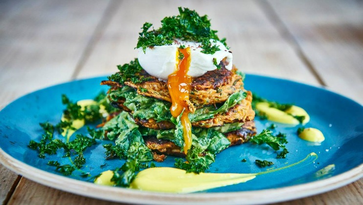 A vibrant blue plate features a stack of savory fritters, topped with a perfectly poached egg with yolk oozing out. Garnished with crispy greens, the dish is surrounded by dollops of creamy yellow sauce. The presentation is visually enticing on a wooden tabletop.