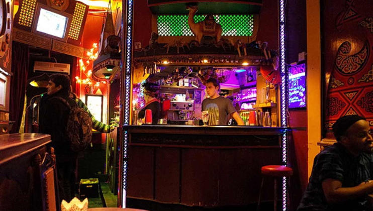 A lively bar with colorful decor, featuring a bartender standing behind the counter. Warm lighting illuminates the space, creating a cozy atmosphere. Patrons are seen enjoying drinks and the vibrant ambiance, with various decorations adorning the walls and ceiling.