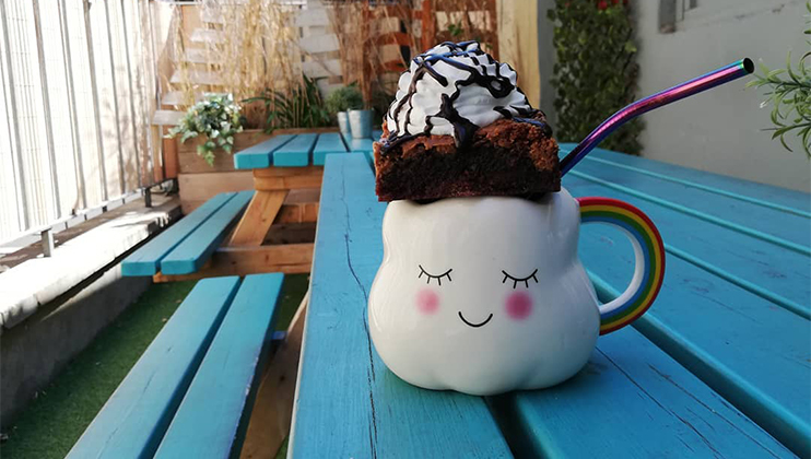 A whimsical cloud-shaped mug with a smiling face, filled with a drink and topped with a brownie, whipped cream, and chocolate drizzle. A rainbow-colored straw sticks out of the mug. The scene is set on a turquoise picnic table in a sunlit outdoor area.