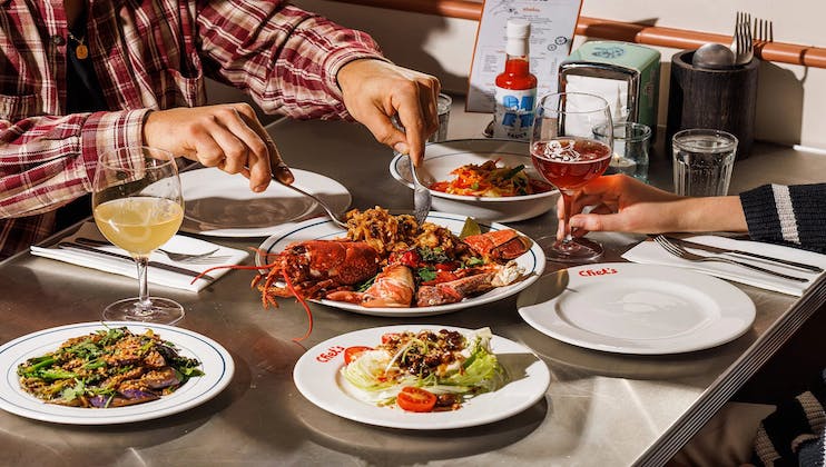 Two people sit at a table laden with various dishes, including lobster, salad, and mixed vegetables. One person is cutting their food, while the other reaches for a drink. There are wine glasses, a menu, and condiments on the table, all set in a cozy restaurant atmosphere.