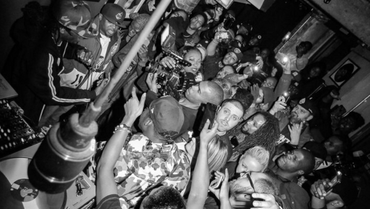 Black and white photo of a crowded room filled with people, many holding cameras and phones. In the center, a person is gesturing with their hand, while others focus their attention on them. The atmosphere appears energetic and lively.