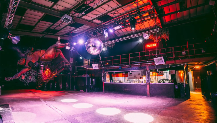 A vibrant nightclub interior with colorful lighting and a large mirror ball hanging from the ceiling. The room features an elevated platform with railings, a bar area at the back, and an art installation of a horse on the left. The space is empty.