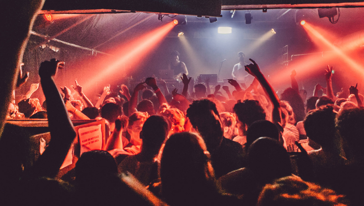 A lively concert scene with a packed audience, hands raised in the air, under vibrant red and orange stage lights. Two performers are visible on stage with musical equipment, creating an energetic and electrifying atmosphere.