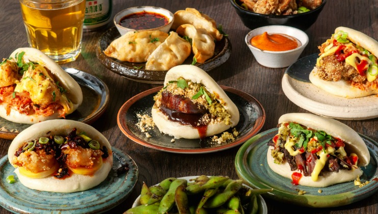 A table spread of Asian-inspired dishes, featuring various steamed buns filled with different ingredients like fried chicken, pork belly, and shrimp. Accompanied by bowls of edamame, dumplings, dipping sauces, and a glass of beer.