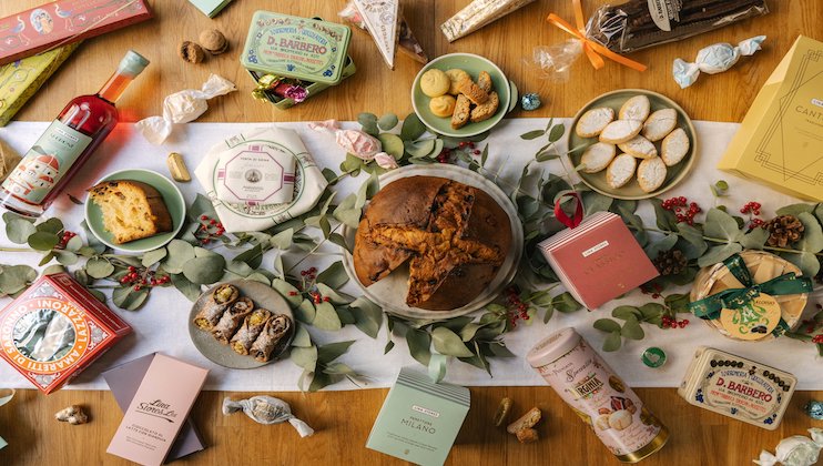 A festive table spread with various packaged and unwrapped pastries, sweets, and bottles. The centerpiece is a large loaf of bread. Leaves and small red berries adorn the table, creating a colorful and celebratory display.