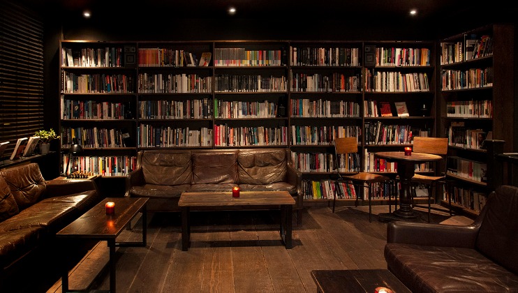 A cozy, dimly-lit library features floor-to-ceiling bookshelves filled with books. The room has brown leather sofas, wooden tables, and chairs. Small red candles on tables add a warm, inviting atmosphere, and a window on the left has closed blinds.