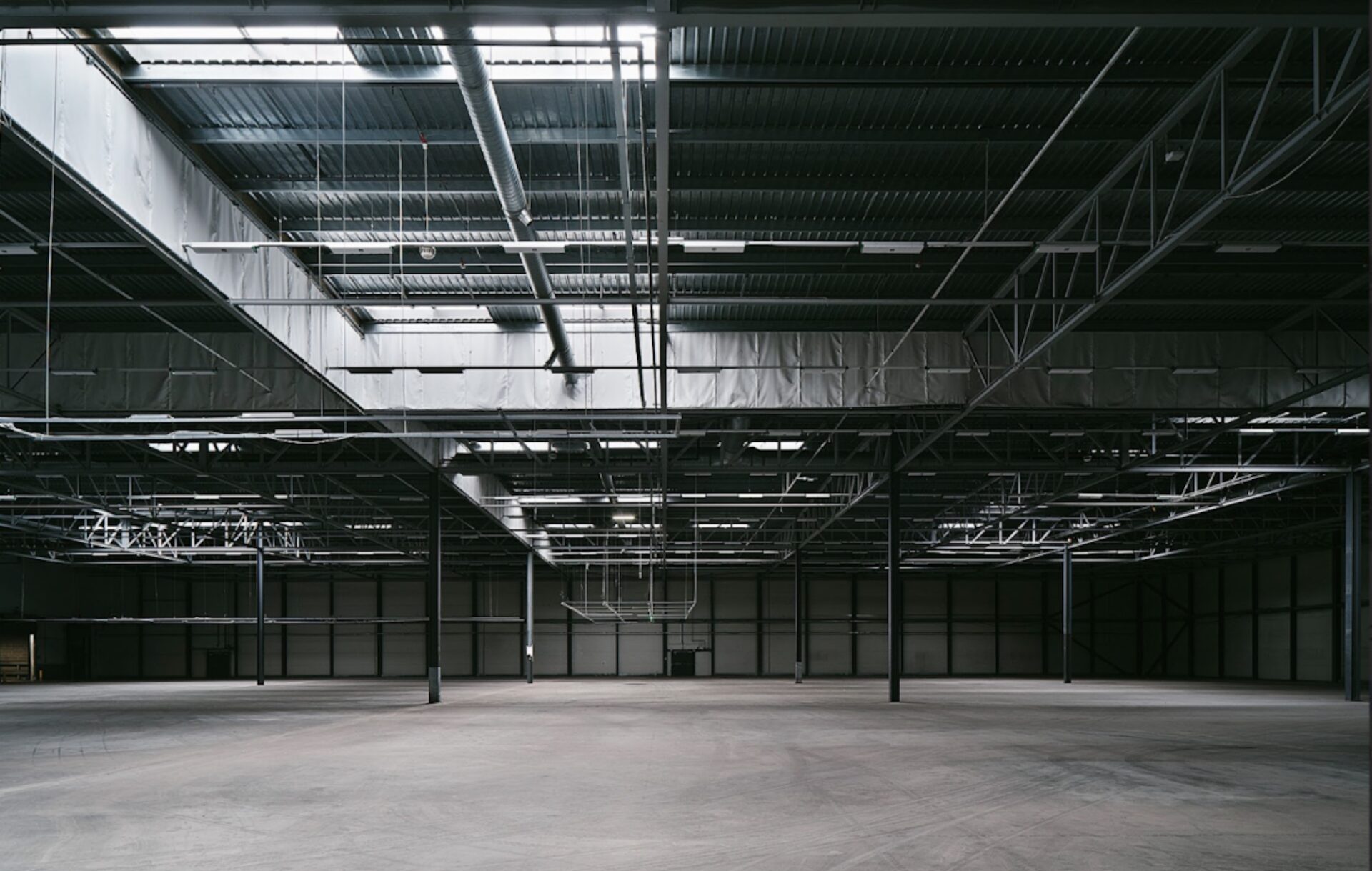 A large, empty industrial warehouse with a high ceiling and exposed metal beams. The floor is smooth concrete, and various pipes and ducts are visible on the ceiling. The space is well-lit from overhead skylights, creating a clean and open atmosphere.