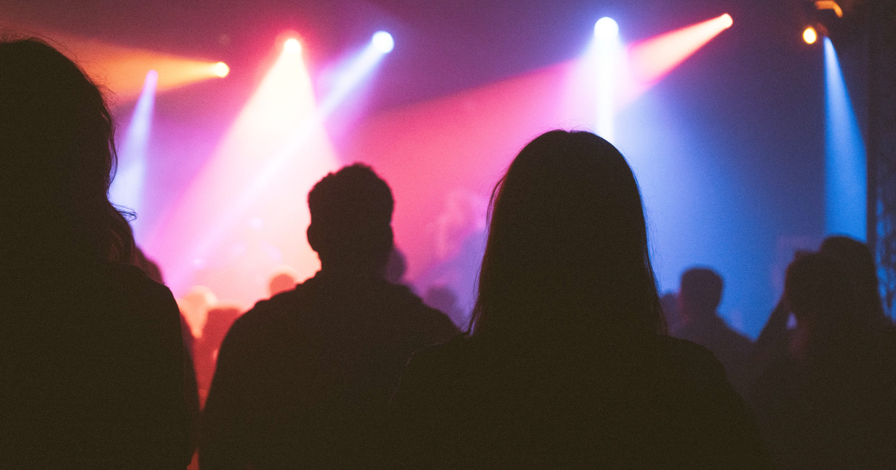 Silhouettes of people standing and facing a vibrant stage with multicolored lights, including pink, blue, and orange hues, creating a lively atmosphere. The figures appear to be watching a performance in a darkened venue.