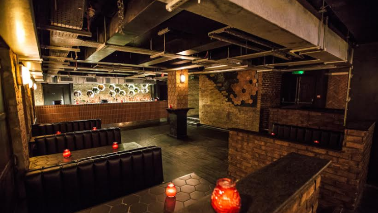 A dimly lit bar interior with exposed ceiling pipes and a geometric honeycomb-like wall design. The seating areas feature black upholstered booths, and small tables with red candle-lit lanterns are placed throughout the space.