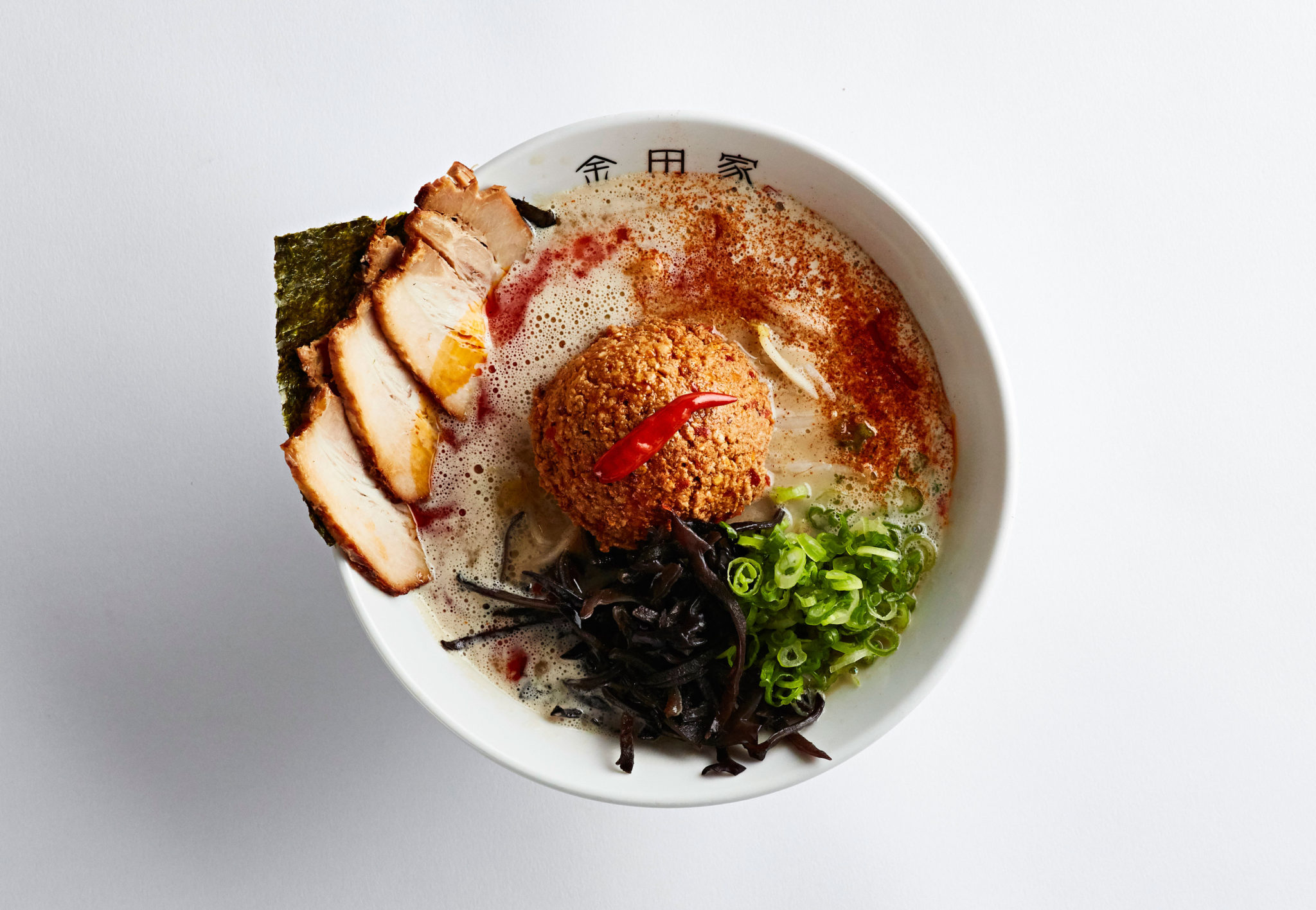 A bowl of ramen featuring a rich broth, slices of pork, green onions, seaweed, black fungus, and a crispy fried ball topped with a small red chili pepper. The ingredients are arranged decoratively, showcasing a variety of textures and colors.